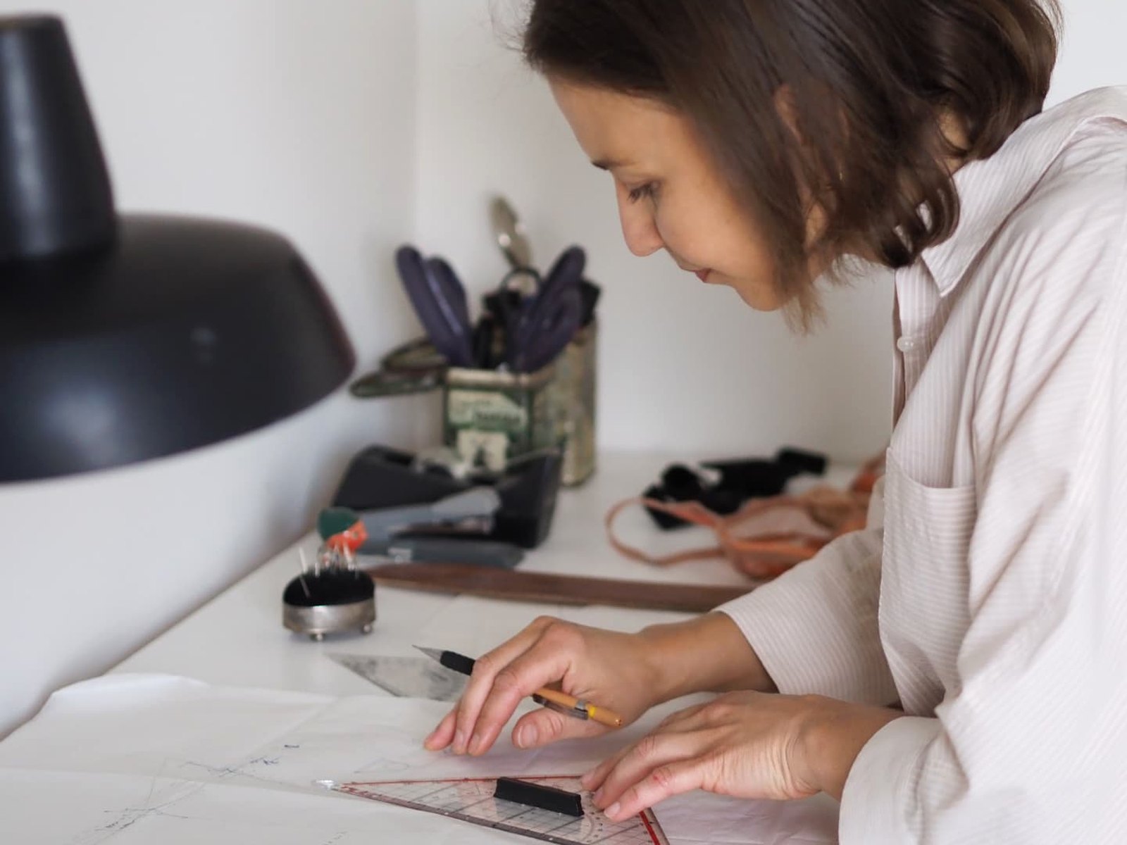 Belgian lingerie designer Els working on the Vicky Baey Essential Tucking Thong pattern at her white studio table, with pattern paper, ruler and pins.